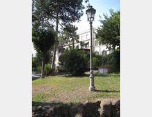 Lampione in ferro in un’area verde con prato, alberi alti e cespugli, delimitata da un muretto in pietra. Sullo sfondo si vede un edificio chiaro a più piani con finestre e balconi, parzialmente nascosto dalla vegetazione.