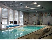 Piscina coperta con grande vasca rettangolare e acqua turchese, illuminata da ampie finestre a tutta altezza su un lato. Pareti decorate con murales di figure atletiche e trampolino alto con piattaforme, con tribune e corrimano lungo il bordo vasca.