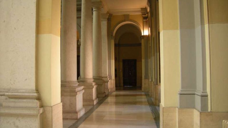 Corridoio interno di un edificio storico con alti pilastri e colonne in pietra lungo il lato sinistro, pareti color crema e pavimento in marmo. In fondo si vede un grande arco e una porta scura, con illuminazione soffusa a parete.