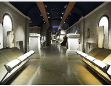 Interno di un corridoio museale lungo e simmetrico con pareti in mattoni e soffitto a volta scuro, illuminato da faretti. Ai lati si vedono basamenti e reperti in pietra, pannelli espositivi inclinati e grandi aperture ad arco incassate nelle pareti.