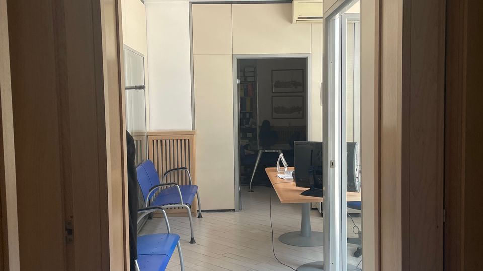 Corridoio interno di un ufficio con pavimento chiaro a listoni e pareti bianche, illuminato da faretti a soffitto. A sinistra ci sono sedute blu in fila, mentre in fondo si vede un’area reception con scrivania e computer, separata da una porta a vetri e con altri ambienti sullo sfondo.