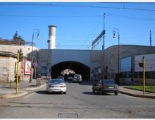 Strada urbana che conduce a un ampio sottopasso in muratura con grande arco centrale, pareti con cartelloni e una ciminiera sullo sfondo. Sopra il tunnel si vedono cavi della linea ferroviaria e lampioni stradali ai lati della carreggiata.
