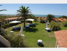 Giardino di un resort sul mare con ampio prato, palme e tavoli apparecchiati con tovaglie bianche sotto ombrelloni. Sullo sfondo si vedono la spiaggia con file di ombrelloni arancioni e, ai lati, edifici bassi con tetti in coppi rossi.