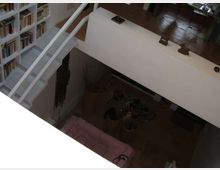 Interno di una casa su due livelli con soppalco e scala bianca; a sinistra c’è una grande libreria a parete e sotto si intravede una zona giorno con divano e vasi decorativi. Pavimento in legno e parapetti bianchi definiscono lo spazio aperto.