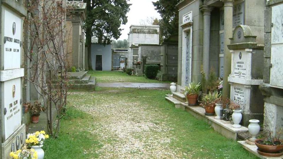 Viale interno di un cimitero con sentiero di ghiaia e prato, affiancato da tombe e cappelle in pietra e marmo con iscrizioni. Ai lati si vedono vasi di fiori e piccoli arbusti, con alberi alti sullo sfondo.