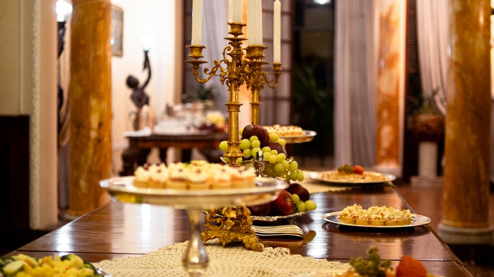 Sala da pranzo elegante con lungo tavolo di legno apparecchiato a buffet, piatti e alzate, e candelabro dorato al centro. L’ambiente è incorniciato da colonne in marmo e grandi finestre con tende chiare sullo sfondo.