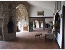 Ampio salone interno con pavimento in cotto e pareti in pietra e intonaco, attraversato da grandi archi in muratura che conducono a corridoi e stanze sullo sfondo. Sulla destra sono presenti specchi e quadri alle pareti e un piccolo salottino con poltrone e tavolino, con piante e decorazioni floreali.