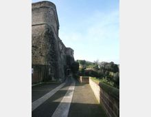 Camminamento lastricato lungo una grande fortificazione in pietra con torre e mura massicce, con un passaggio ad arco in fondo. A destra un muretto di mattoni affaccia su una valle verde con alberi e alcune case in lontananza.