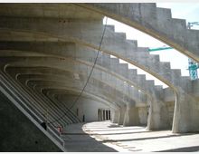 Interno di una grande struttura in cemento armato con una serie di archi e travi curve ripetute che formano un corridoio coperto. Sulla destra si vede una scalinata integrata nella parete e, sullo sfondo, aperture verso l’esterno con luce naturale.