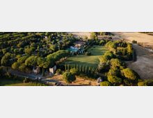 Veduta aerea di una tenuta di campagna con ampio prato centrale, filari di cipressi e boschi di pini mediterranei, con alcuni edifici in pietra e una piscina vicino alla villa principale. Ai margini si vedono strade sterrate e campi coltivati.