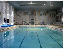 Interno di una piscina coperta con vasca rettangolare e corsie segnate sul fondo, circondata da bordo in piastrelle e attrezzature da nuoto. Sulle pareti alte sono presenti grandi murales con figure atletiche, gradinate laterali e ampie finestre che illuminano l’ambiente.