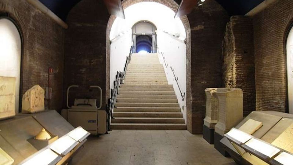 Interno di uno spazio museale in mattoni con soffitto a volta e ampio arco centrale, dominato da una scalinata che conduce a un’uscita illuminata. Ai lati della sala ci sono teche e basamenti con reperti esposti lungo le pareti.
