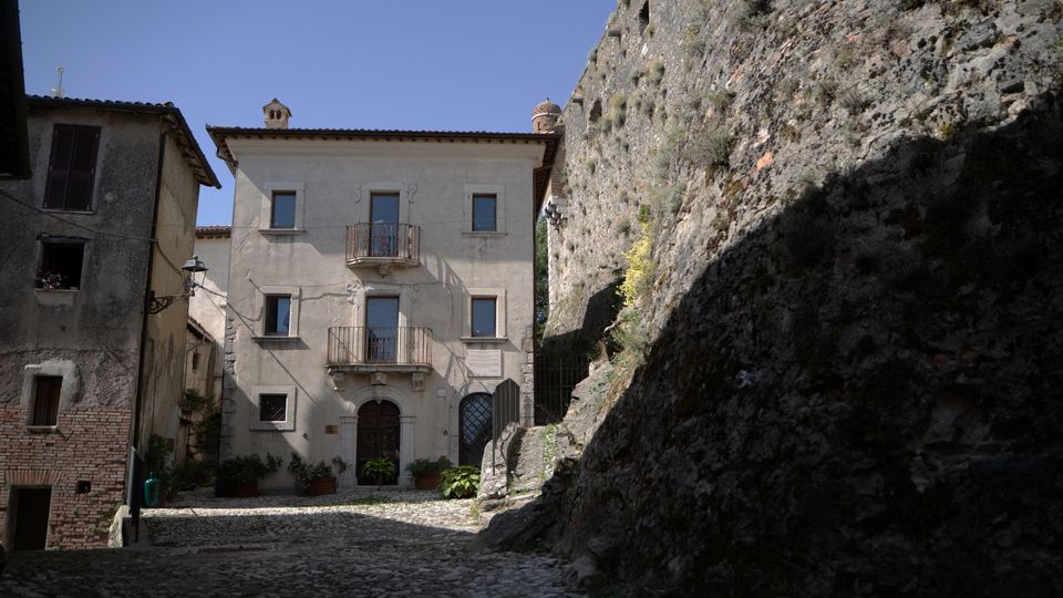 Piazzetta acciottolata in un borgo storico, con edificio a tre piani intonacato chiaro e balconi in ferro battuto al centro. A destra si alza una parete rocciosa ripida con vegetazione sparsa, mentre a sinistra si vedono case in pietra con finestre e una lanterna a muro.