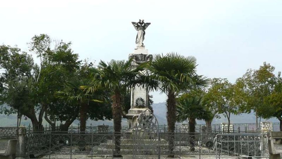 Monumento in pietra con statua sulla sommità, al centro di una piccola area recintata con cancellata in ferro battuto e pavimentazione in ciottoli. Intorno ci sono palme e altri alberi, con vista sul lago e una balaustra sullo sfondo.