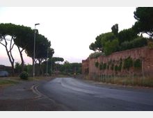 Strada asfaltata in lieve curva con marciapiede e lampioni, fiancheggiata da alti pini mediterranei. Sulla destra corre un lungo muro in mattoni coperto in parte da vegetazione, con recinzione alla base.