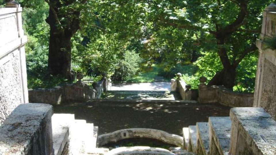 Scalinata in pietra che scende in un’area verde, con balaustre e muretti laterali in muratura. Grandi alberi con chioma fitta ombreggiano il percorso, che conduce a un vialetto luminoso in fondo.