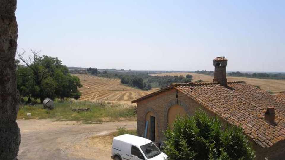 Casale rurale in mattoni con tetto in coppi e camino, affacciato su una strada sterrata con un furgone parcheggiato. Sullo sfondo si estendono campi coltivati ondulati e alberi sotto un cielo sereno.