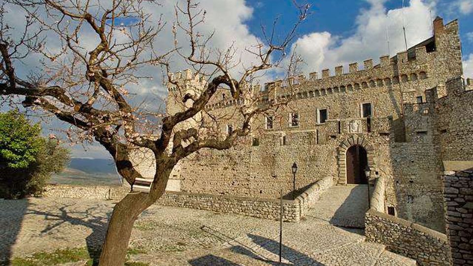 Castello in pietra con merlature e mura fortificate, con ingresso ad arco raggiunto da una rampa su pavimentazione in ciottoli. In primo piano un albero spoglio e un lampione, con cielo parzialmente nuvoloso sullo sfondo.