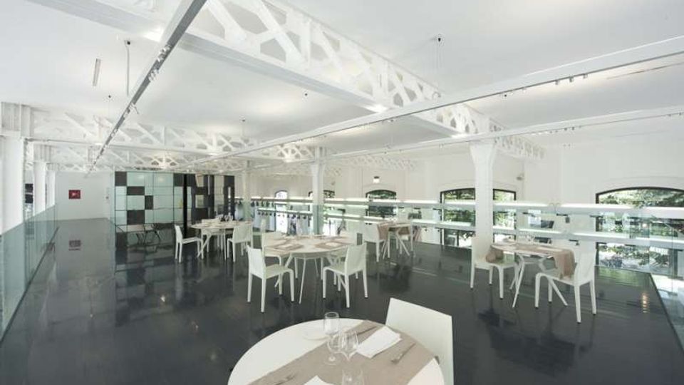 Interno di un ristorante moderno e luminoso con pavimento scuro lucido, tavoli rotondi con sedie bianche e soppalco con ringhiera in vetro. Soffitto alto con travi metalliche bianche a vista e grandi finestre ad arco lungo le pareti.