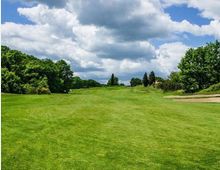 Ampio campo da golf con fairway erboso in leggera salita, circondato da alberi e cespugli ai lati. A destra si vede un bunker di sabbia, con cielo azzurro e grandi nuvole sullo sfondo.