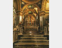 Interno di una chiesa barocca con navata riccamente decorata, archi e soffitto a volta affrescati e dorati. In fondo si vede l’altare rialzato con gradini in marmo, balaustre ornate e organo a canne incorniciato da elementi scultorei.