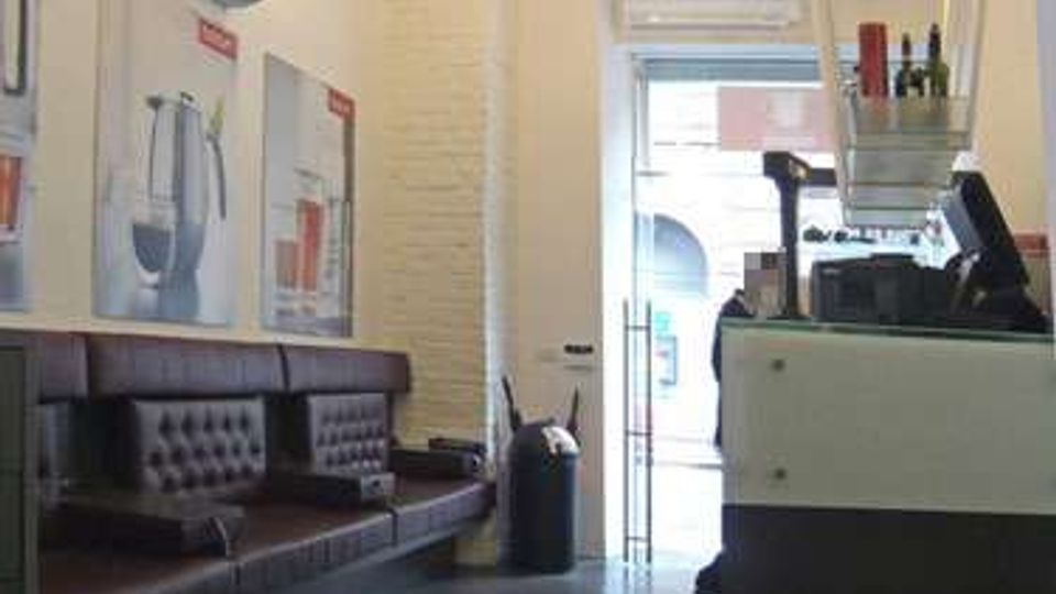 Interno di un piccolo bar o caffetteria con soffitto alto, pareti chiare e luci a sospensione. A sinistra ci sono divanetti in pelle scura lungo la parete con quadri e un condizionatore, mentre a destra si vede il bancone con mensole e attrezzature; in fondo una porta a vetri dà sulla strada.
