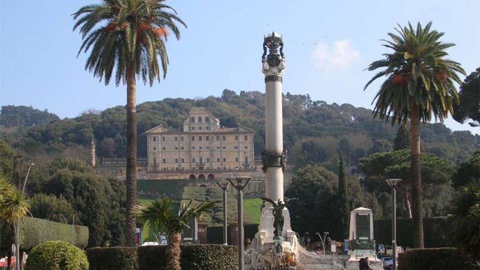 Piazza con giardini curati e alte palme, con una colonna monumentale e fontana al centro. Sullo sfondo, su una collina boscosa, si vede un grande edificio storico in stile palazzo.
