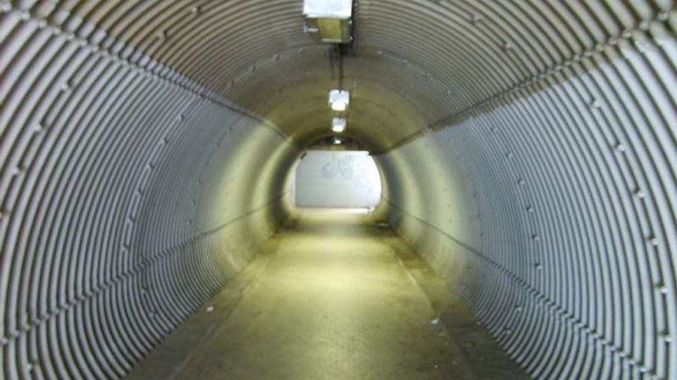 Sottopassaggio pedonale a forma di tunnel con pareti e soffitto in pannelli metallici ondulati, illuminato da lampade rettangolari a soffitto. Il pavimento asfaltato conduce verso un’uscita luminosa sul fondo.