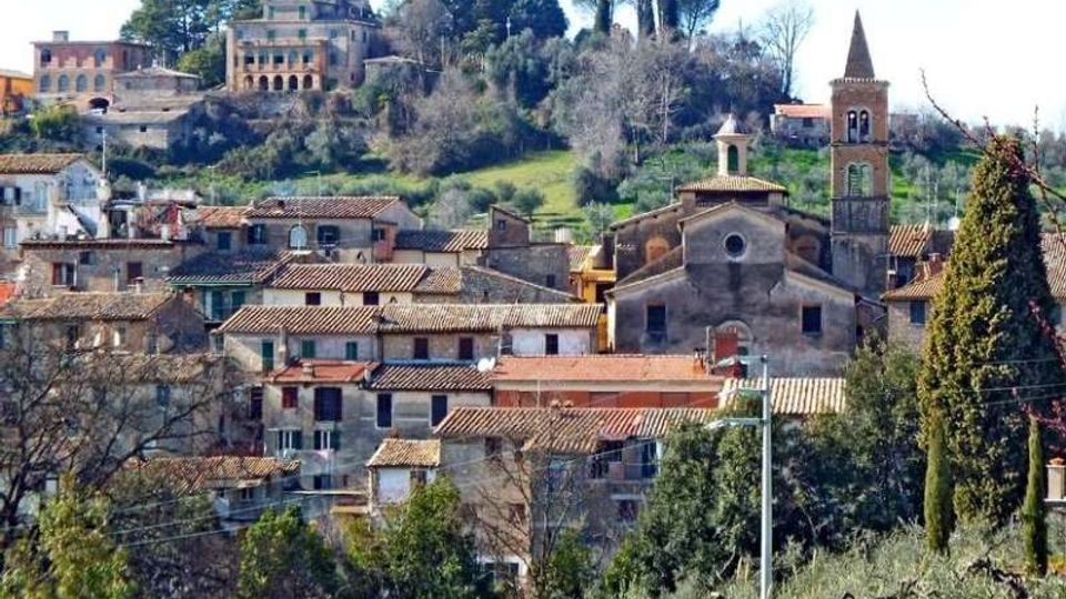 Veduta panoramica di un borgo collinare con case in pietra dai tetti in coppi. In primo piano si distinguono una chiesa con facciata semplice e campanile in mattoni, mentre sullo sfondo si vedono alberi e una grande villa storica sulla sommità della collina.