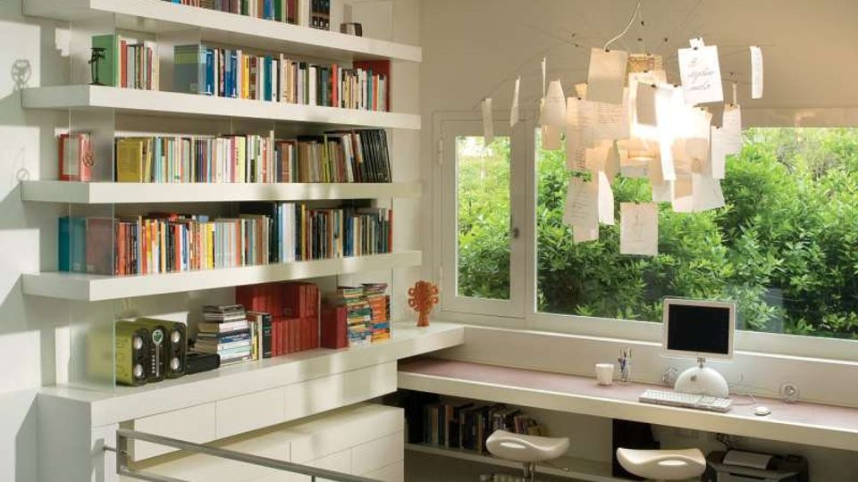 Interno di uno studio domestico moderno con pavimento in legno, libreria a parete e lunga scrivania sotto una grande finestra affacciata sul verde. Il soffitto ha lucernari a fasce e una lampada a sospensione con fogli appesi, con ringhiera metallica lungo il bordo del soppalco.
