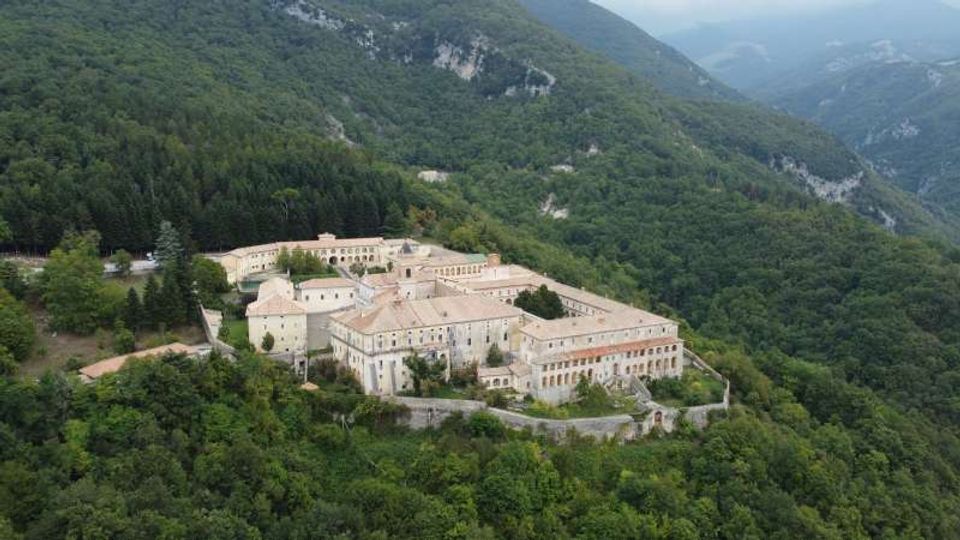 Veduta aerea di un grande complesso monastico in pietra con tetti in laterizio, disposto attorno a cortili e racchiuso da mura. L’edificio sorge su un pendio montano ed è circondato da fitti boschi, con vallate e montagne sullo sfondo.