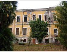 Facciata di un edificio storico a due piani intonacato di giallo, con finestre rettangolari e ampie zone di intonaco scrostato; al centro una grande edera ricopre parte del muro. Davanti si vede un cortile erboso con un piccolo elemento in pietra e vegetazione alta ai lati.