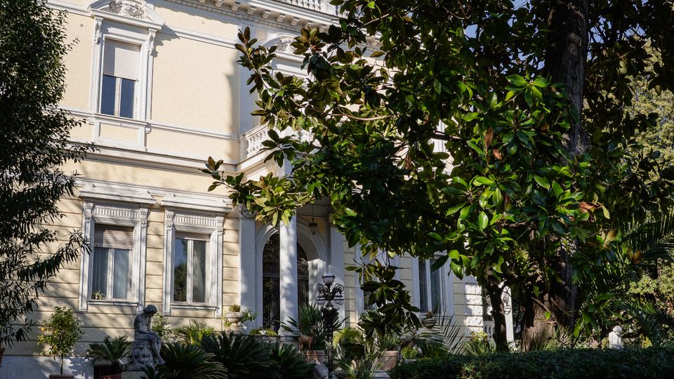 Facciata di una villa storica color crema con finestre incorniciate e balconata in sommità, parzialmente coperta da grandi alberi. In primo piano un vialetto nel giardino con siepi, vasi e una piccola statua vicino all’ingresso con portico.