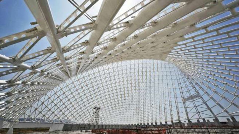 Interno di una grande struttura architettonica moderna con copertura a griglia di travi metalliche bianche e pannelli trasparenti, che forma una volta curva. Ampie aperture lasciano vedere il cielo e, sullo sfondo, una sagoma urbana con gru e edifici.