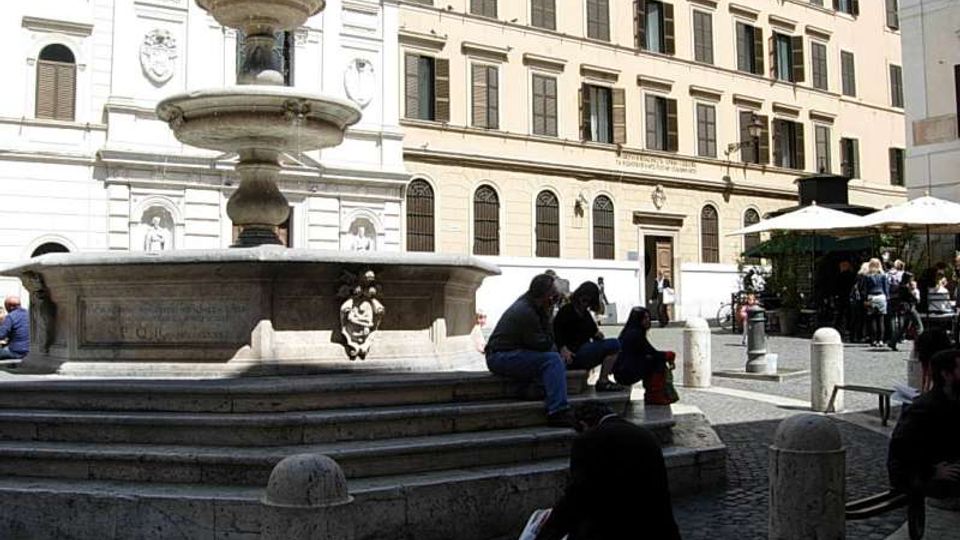 Fontana in pietra a più vasche al centro di una piazza lastricata, circondata da palazzi storici con finestre e persiane. Intorno si vedono dissuasori in pietra, panchine e un’area con tavolini all’aperto sotto grandi ombrelloni.