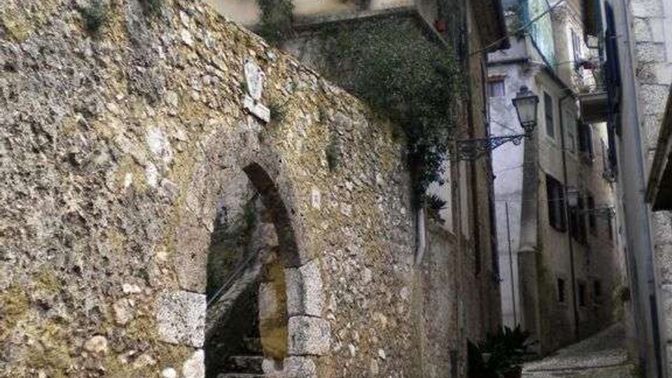 Vicolo stretto in un borgo storico, con pavimentazione in ciottoli e alti muri in pietra. A sinistra si vede un arco con una scalinata che sale verso una casa con balcone e piante, mentre sul lato destro ci sono edifici in muratura e un lampione a parete.
