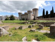 Castello medievale in pietra con quattro torri cilindriche merlate, visto da un’area archeologica con muretti e resti in muratura tra erba e sentieri. Sullo sfondo si notano cipressi e alcuni edifici moderni sotto un cielo parzialmente nuvoloso.