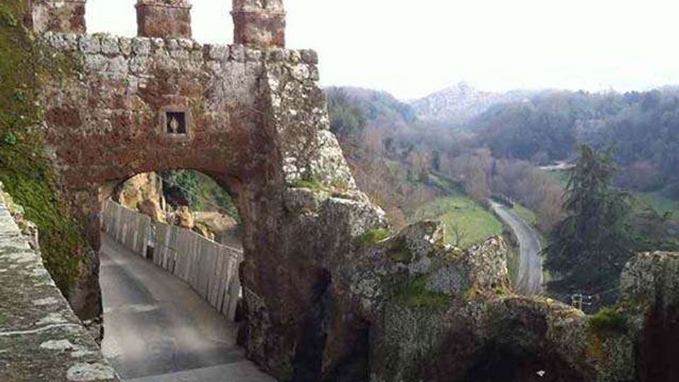 Porta fortificata in pietra con merli e grande arco che scavalca una strada stretta, affiancata da muri rocciosi. Sullo sfondo si vede una valle collinare con boschi e una strada che curva tra i pendii.