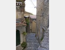 Stretta viuzza in discesa lastricata in pietra, tra edifici in muratura e pietra con tetti in coppi. Un lampione a parete e cavi sospesi attraversano il vicolo, con piccoli ingressi e balconcini affacciati sulla strada.