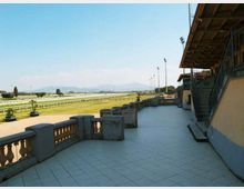 Terrazza pavimentata con balaustra in pietra accanto a una tribuna coperta con scale esterne e ringhiere metalliche. Sullo sfondo si vedono i rettilinei di una pista ippica con prato, pali dell’illuminazione e colline lontane sotto cielo sereno.