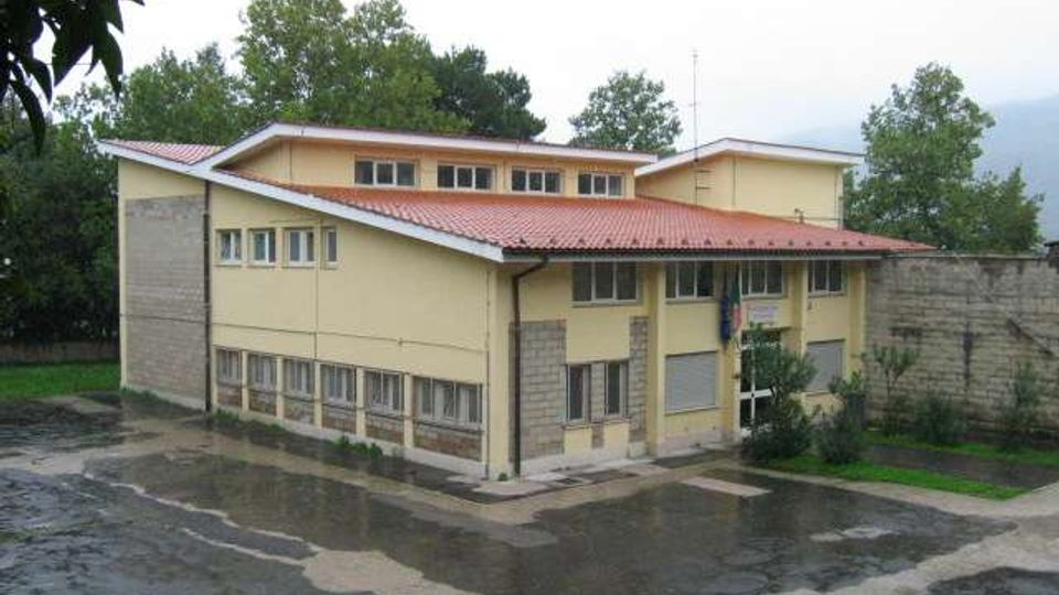 Edificio pubblico a due piani con facciata giallo chiaro e tetto in lamiera rossa, con molte finestre e ingresso centrale. Davanti si vede un cortile asfaltato bagnato dalla pioggia, con alberi sullo sfondo e un muro in cemento sul lato destro.