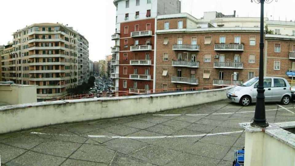 Terrazza urbana in cemento con pavimentazione a grandi lastre e parapetto basso, affacciata su una strada tra alti edifici residenziali in mattoni con balconi. In primo piano si vedono un lampione e un’auto parcheggiata vicino al bordo della terrazza.