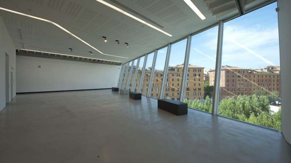 Ampio interno moderno e vuoto, con pavimento in cemento e soffitto con lunghe luci lineari, affacciato su una parete vetrata inclinata a tutta altezza. Attraverso le finestre si vedono edifici residenziali e alberi all’esterno.
