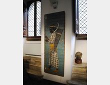 Interno di museo o galleria con pareti bianche e grandi finestre ad arco con vetri a losanghe e scuri in legno. Al centro è esposto un pannello verticale decorato in stile antico su supporto azzurro, con una piccola scultura su basamento accanto e gradini in pietra sul lato.