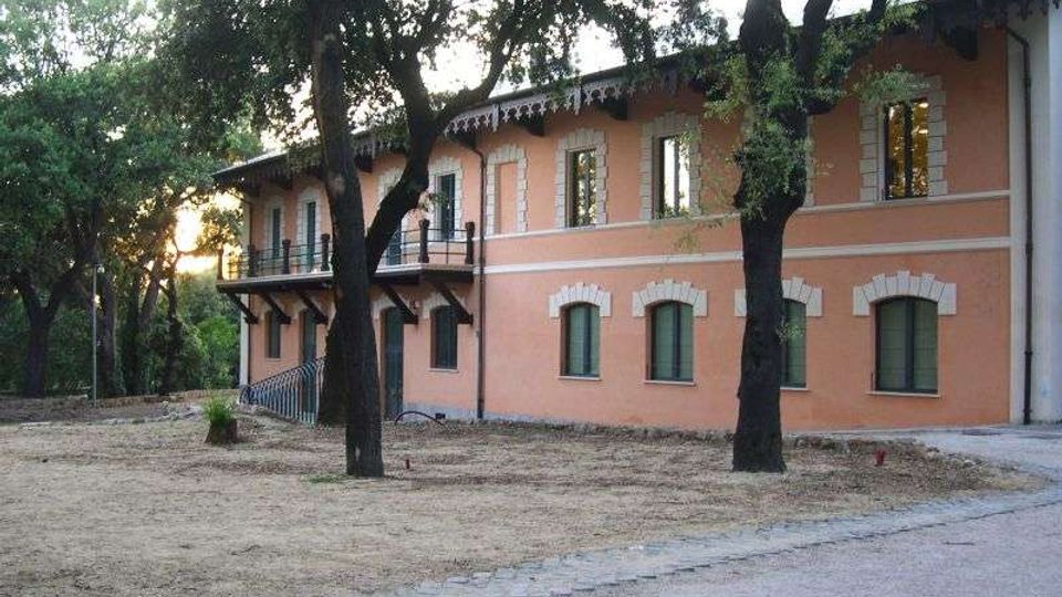 Edificio a due piani color salmone con cornici bianche attorno alle finestre e balconata esterna, situato in un’area alberata. In primo piano si vedono alberi ad alto fusto e un vialetto di ghiaia che costeggia la facciata.