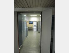 Corridoio interno di una struttura ospedaliera o clinica, con pavimento in linoleum grigio, pareti bianche e armadi metallici lungo il lato sinistro. In fondo si vedono una barella e una porta aperta verso un’altra stanza illuminata da lampade fluorescenti.