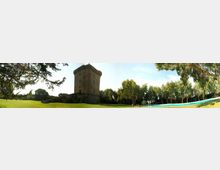 Torre in pietra medievale a pianta quadrata su un terrapieno, circondata da un ampio prato verde e da alberi. In primo piano si vede una lunga piscina rettangolare con bordo azzurro sotto un cielo sereno.