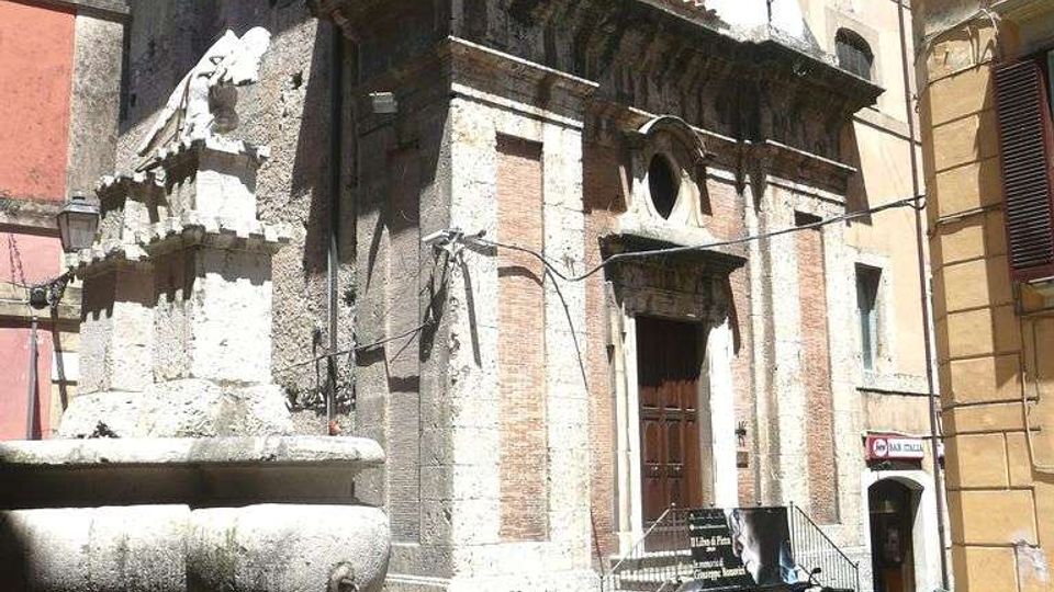 Piccola piazza urbana con una fontana in pietra in primo piano e una chiesa storica sullo sfondo, con facciata in pietra e mattoni, portale centrale e frontone curvilineo. Intorno si vedono edifici alti e ravvicinati lungo una strada stretta, con alcune moto parcheggiate vicino all’ingresso.