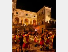 Palazzo storico illuminato di notte con loggiato ad archi e ampia scalinata centrale che conduce al piano superiore. In primo piano si vede una piazza lastricata in pietra, circondata da facciate antiche.