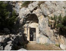 Ingresso di una grotta scavata in una parete rocciosa, con cancello in ferro battuto e una croce fissata alla roccia accanto. L’area circostante è un sentiero sterrato tra massi, vegetazione bassa e pini sulle rocce.